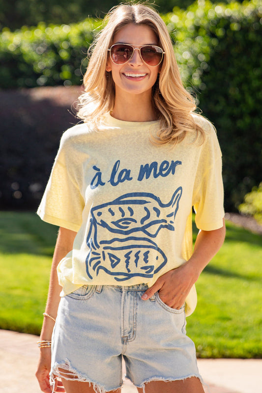 Woman wearing a yellow t-shirt with blue graphics and text, standing outdoors.