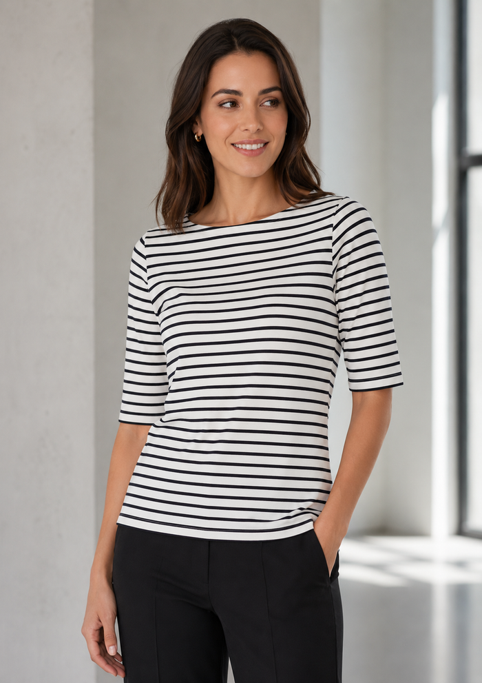 A woman wearing a black and white horizontally striped boat-neck top with elbow-length sleeves, paired with black trousers, standing in a brightly lit modern interior.